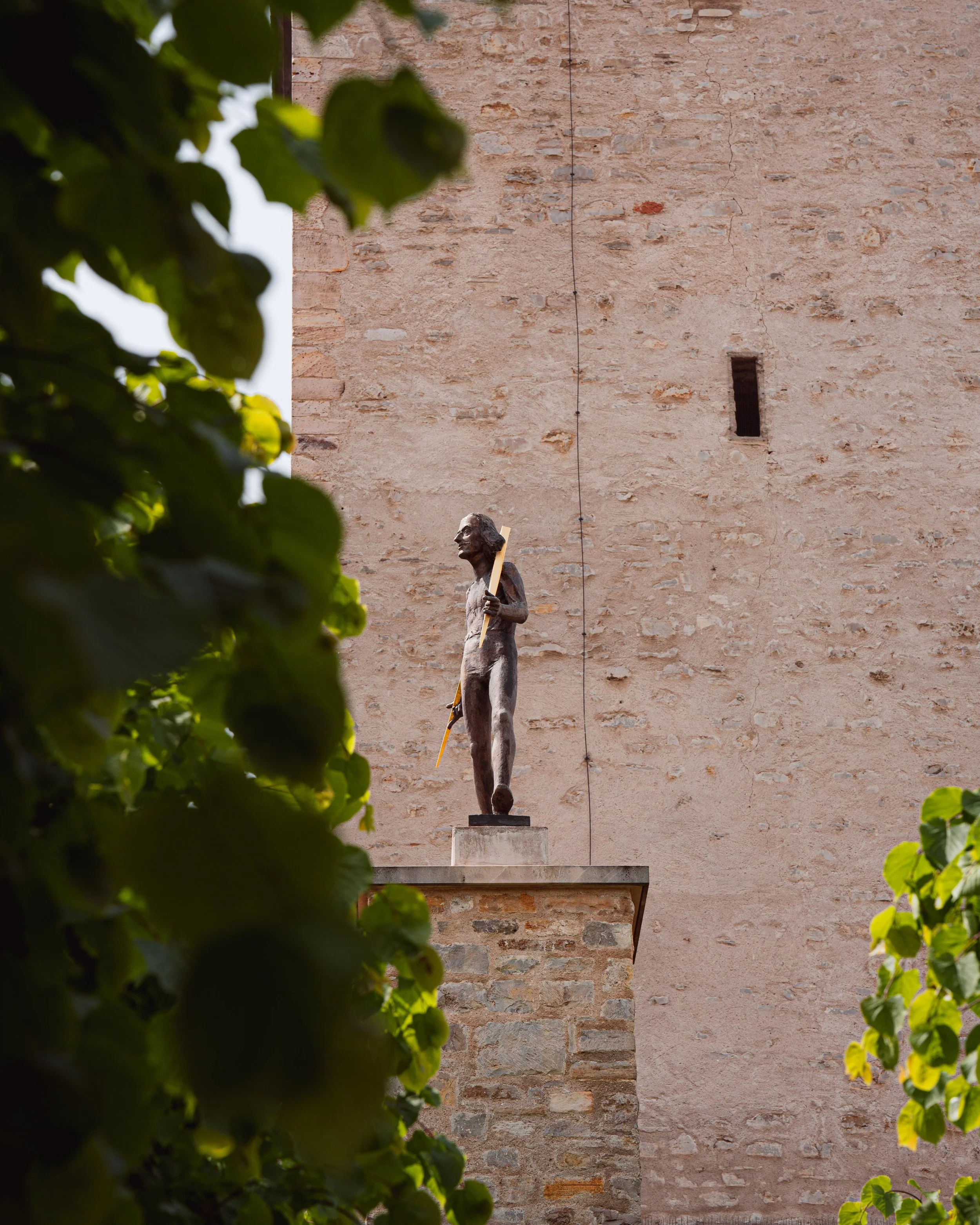 Eine Skulptur eines Mannes, der eine lange Stange hält, vor einer alten Steinmauer. Der obere Bereich der Mauer ist sichtbar, eingeritzte Linien und ein kleiner Schlitz sind vorhanden, umgeben von grünem Laub.