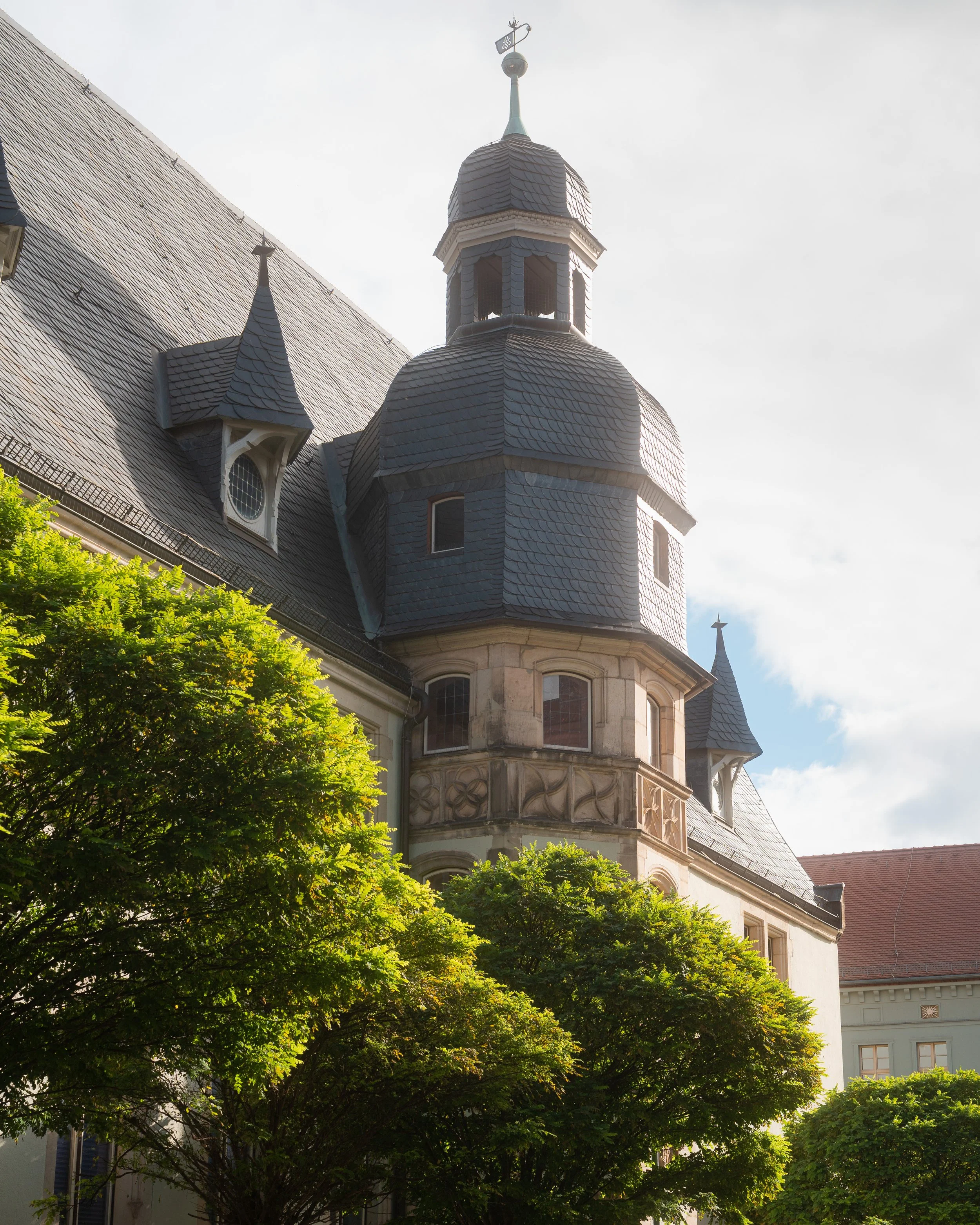Gotischer Turm mit Schindeldach und Wetterfahne auf dem Dach, hinter grünem Baum. Auffällige Architektur und Blautöne auf dem Gebäude, darunter mehrere Fenster und eine Kombination aus Stein- und Holzstrukturen.