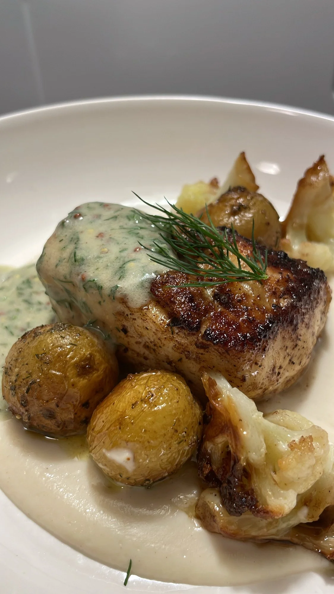 A plated dish featuring a piece of grilled chicken topped with herb sauce, roasted potatoes, cauliflower, and garnished with fresh dill.