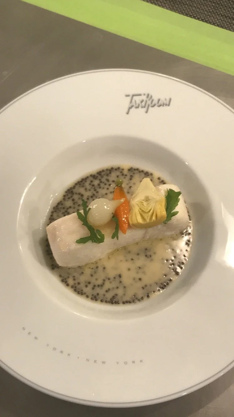 A gourmet dish featuring a piece of white fish atop a bed of black caviar and creamy sauce, garnished with a small onion, carrot, a dollop of butter, and fresh herbs on a white plate from Tartare restaurant in New York.