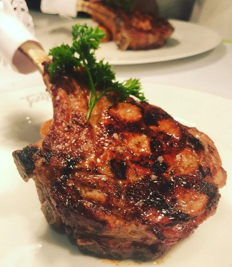 Close-up of a grilled beef steak with herbs on top, served on a white plate, with another steak in the background.