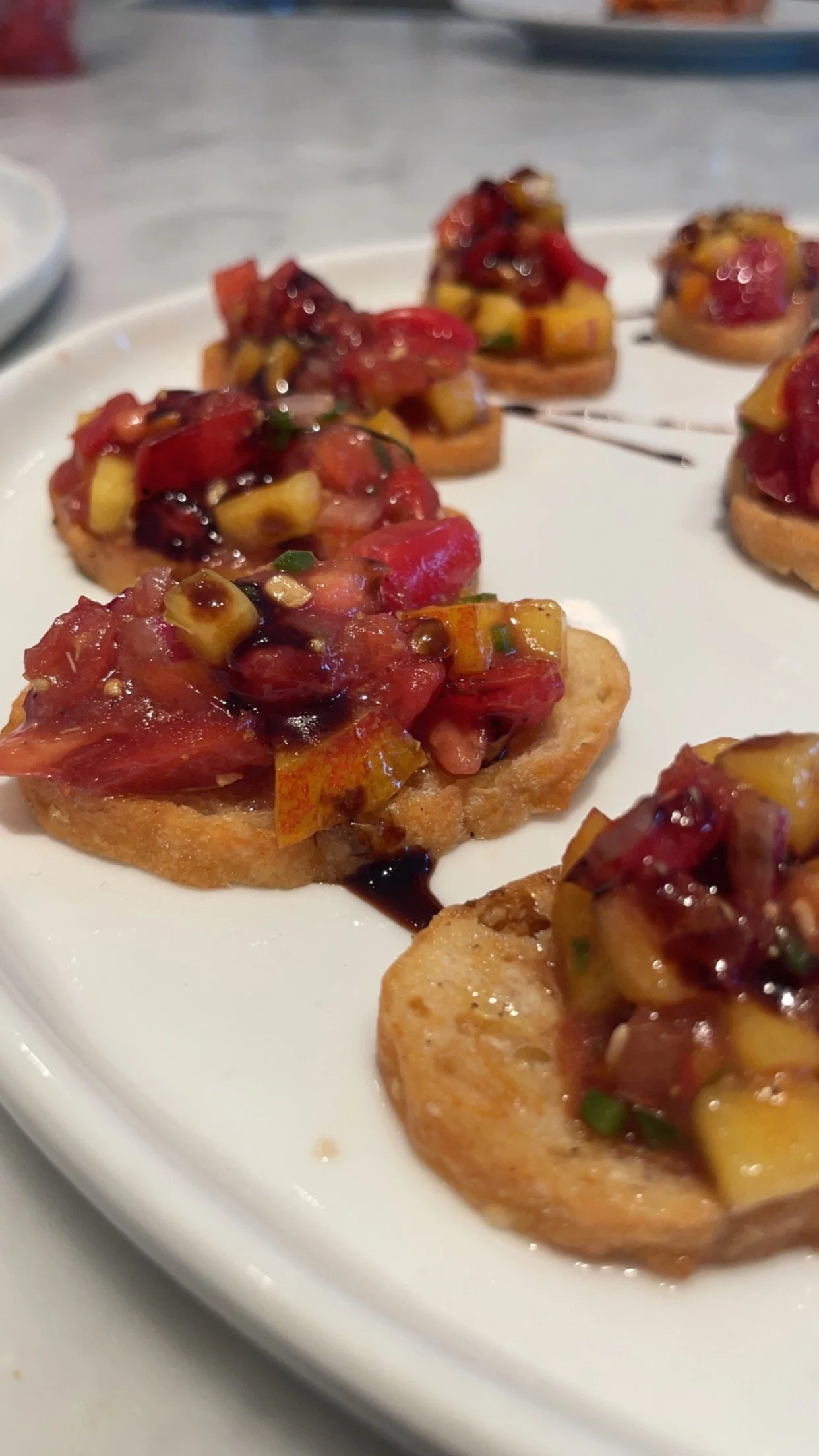Close-up of toasted bread topped with diced tomatoes, mango, and herbs, drizzled with balsamic glaze, on a white plate.