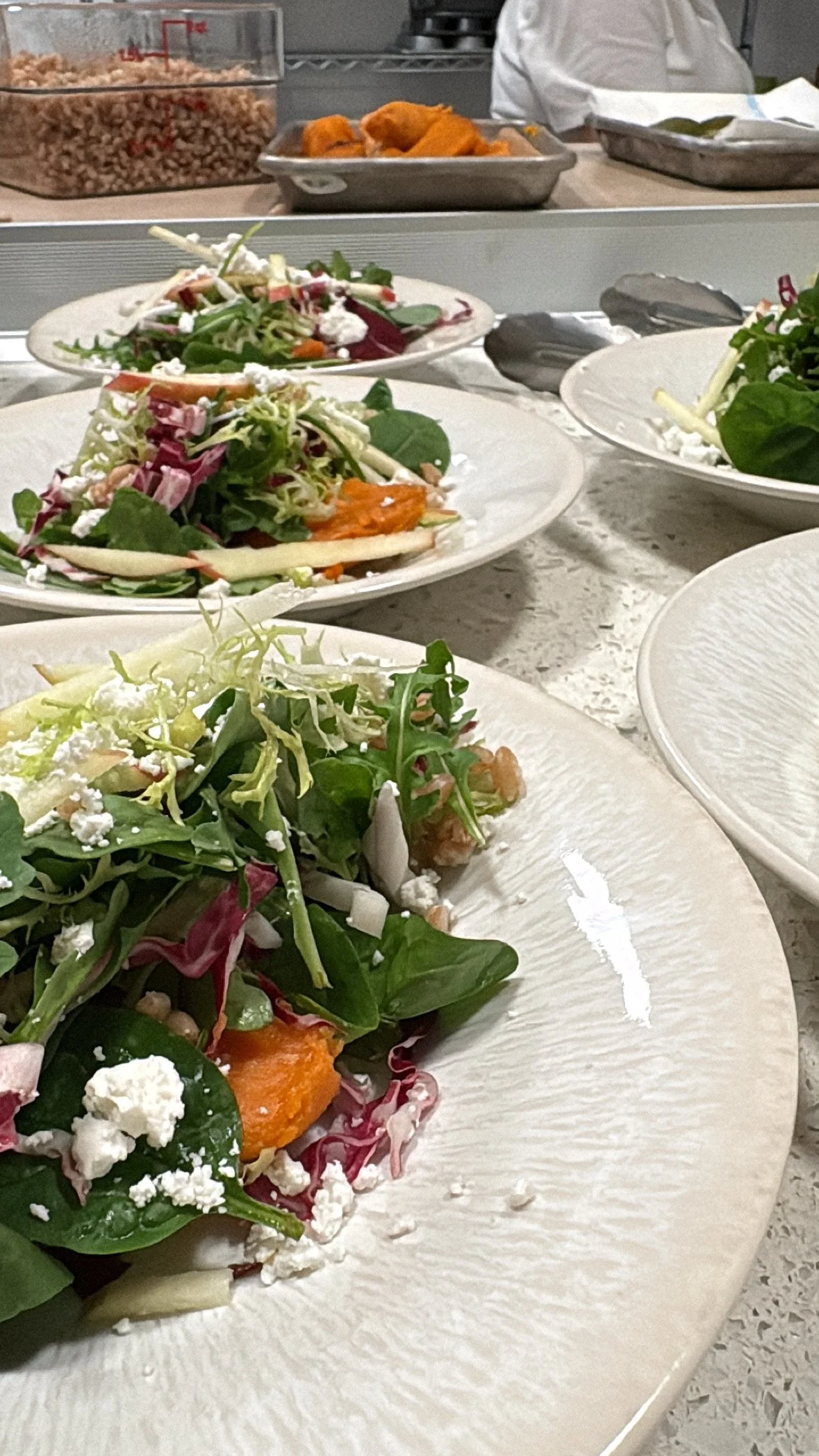 Multiple plates of fresh mixed green salads with vegetables, cheese, and possibly grains, in a professional kitchen environment with food prep containers and kitchen staff in the background.