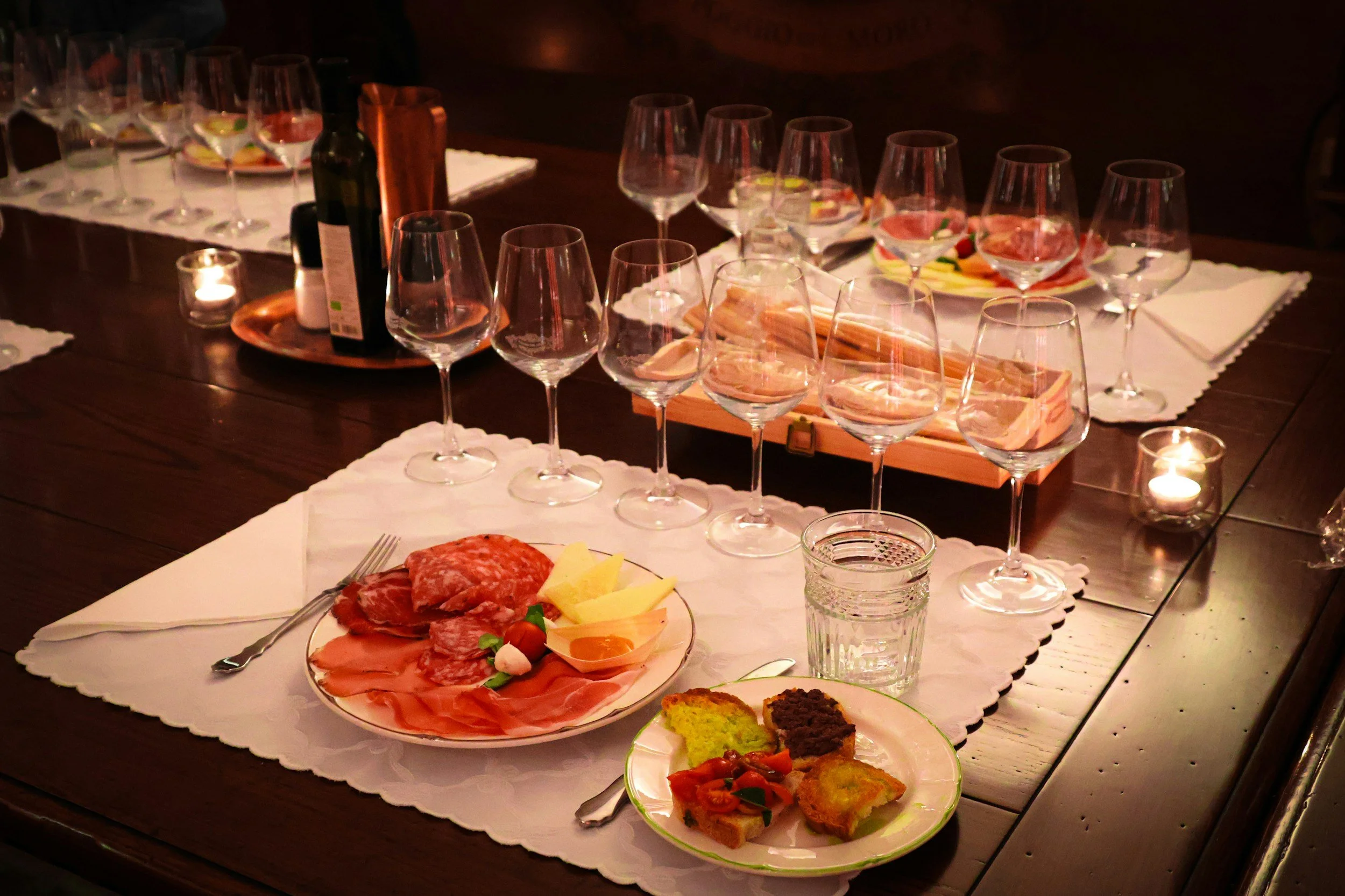 A table set with empty wine glasses, plates of assorted cheeses and meats, and small appetizers, lit by candlelight.