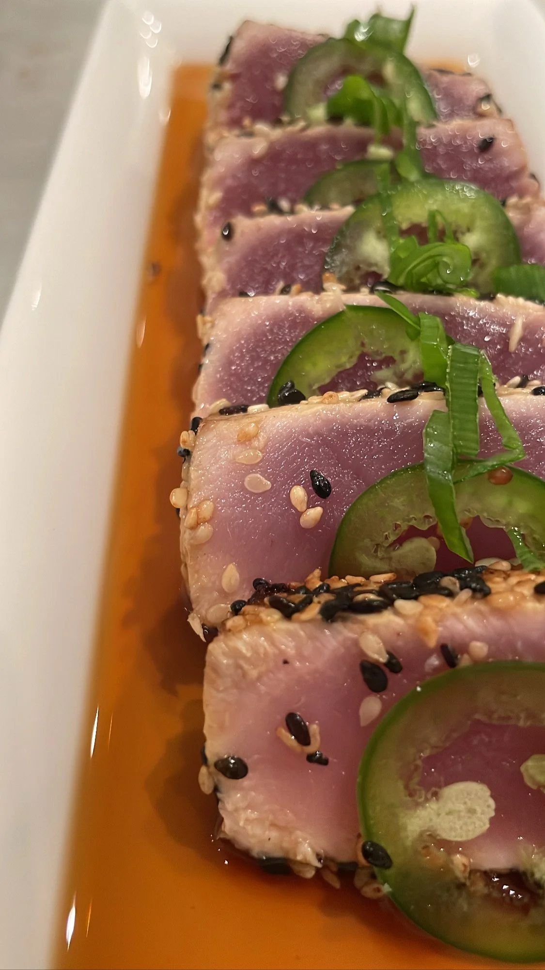 Slices of tuna tuna topped with cucumber slices and green onion, sprinkled with sesame seeds and black sesame seeds, served in a light soy sauce
