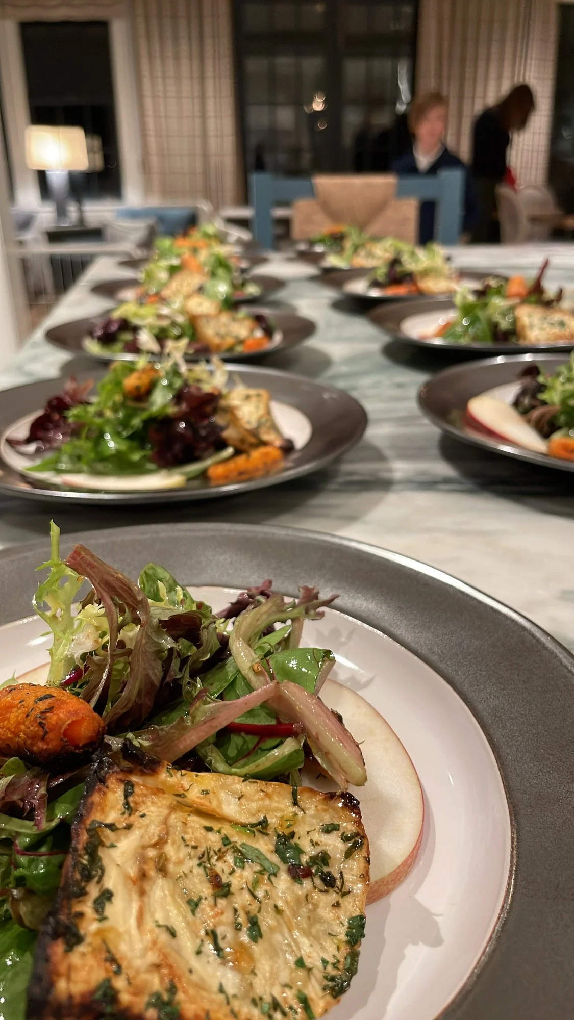 Close-up of plated salads with grilled vegetables, greens, and garnishes on a long dining table in a cozy dining room.