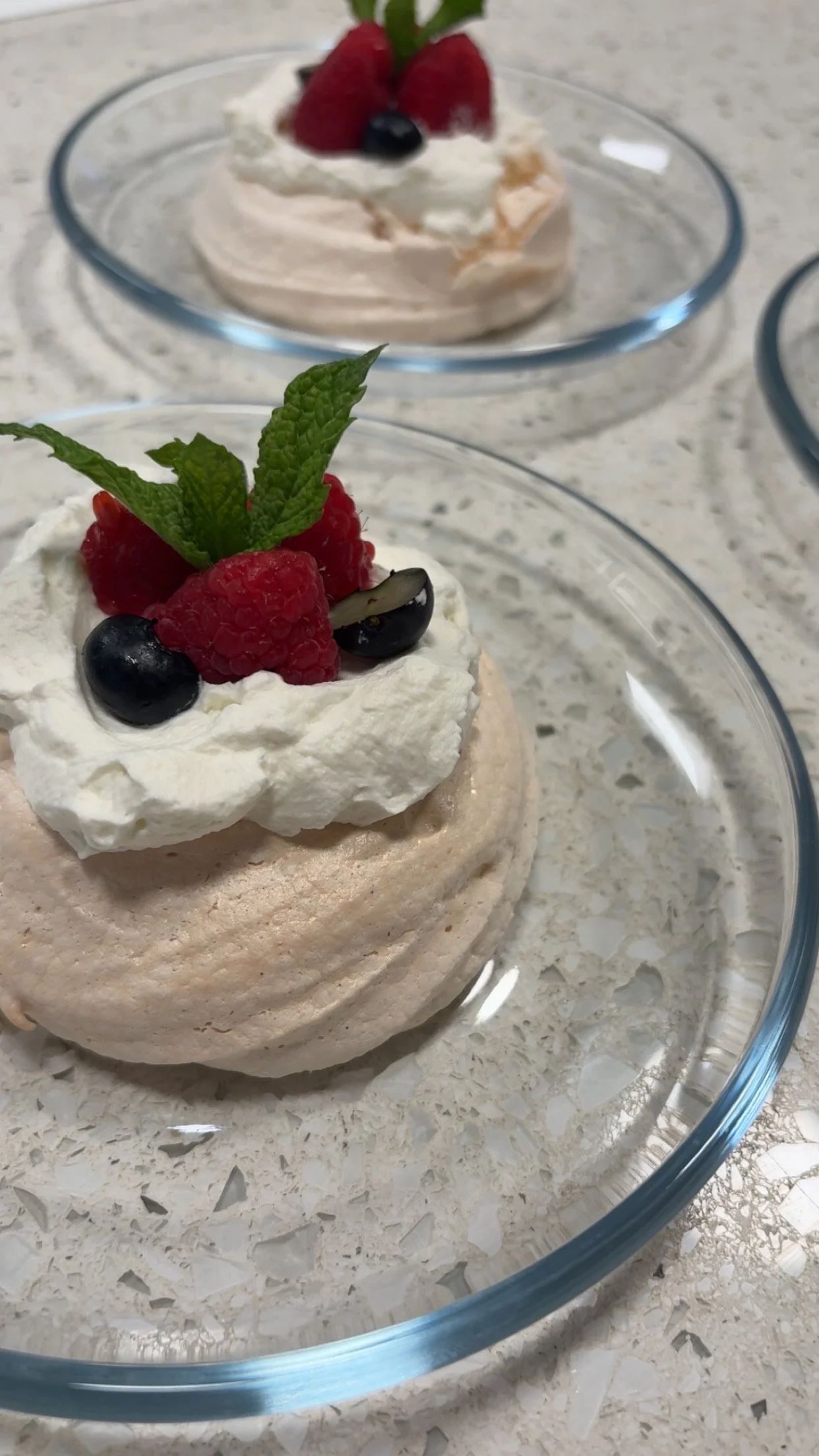 A dessert with whipped cream, raspberries, blueberries, and a mint leaf on top, served on a glass plate.