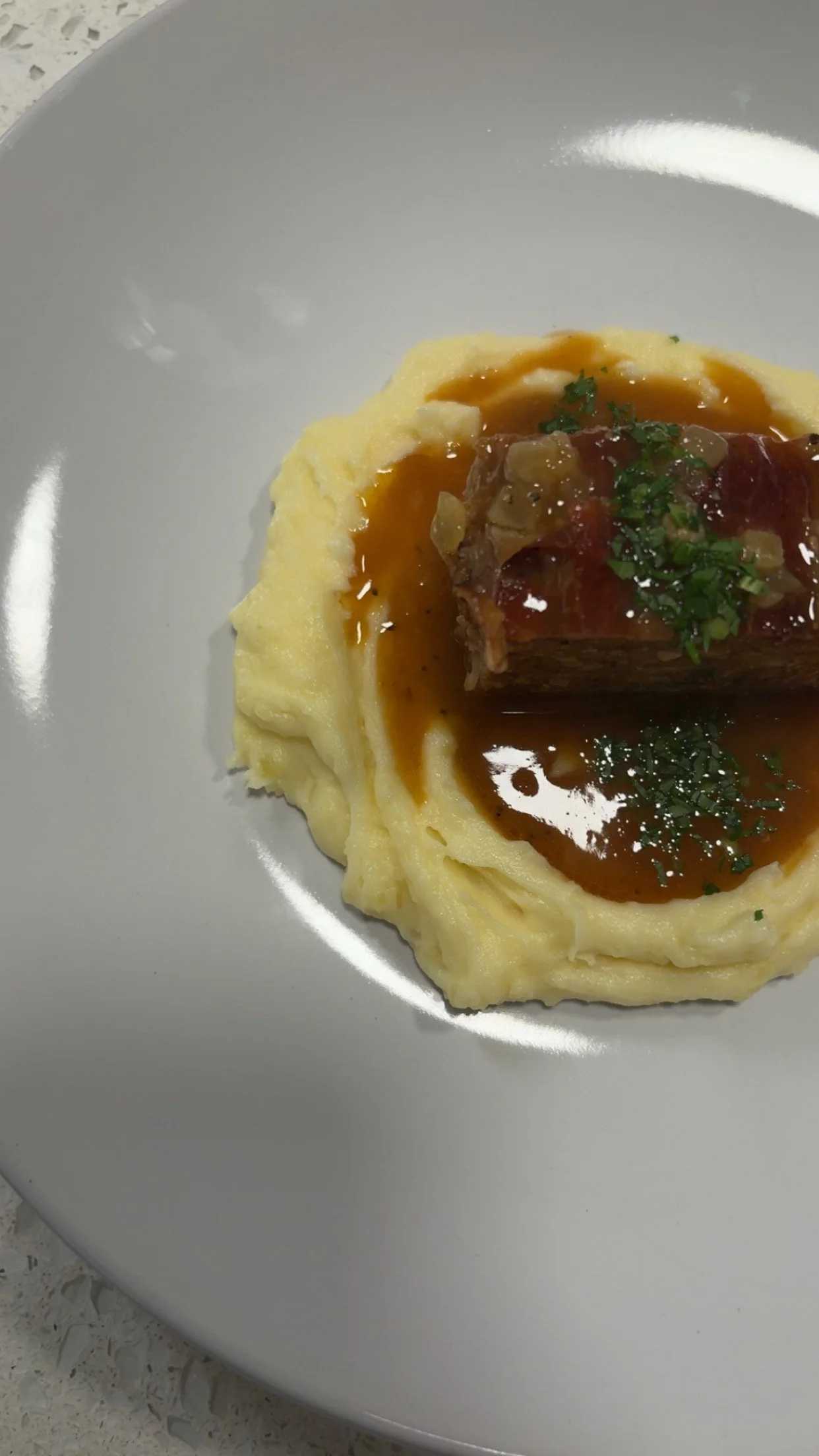 A plate with mashed potatoes topped with a slice of meatloaf, garnished with parsley, and drizzled with gravy.