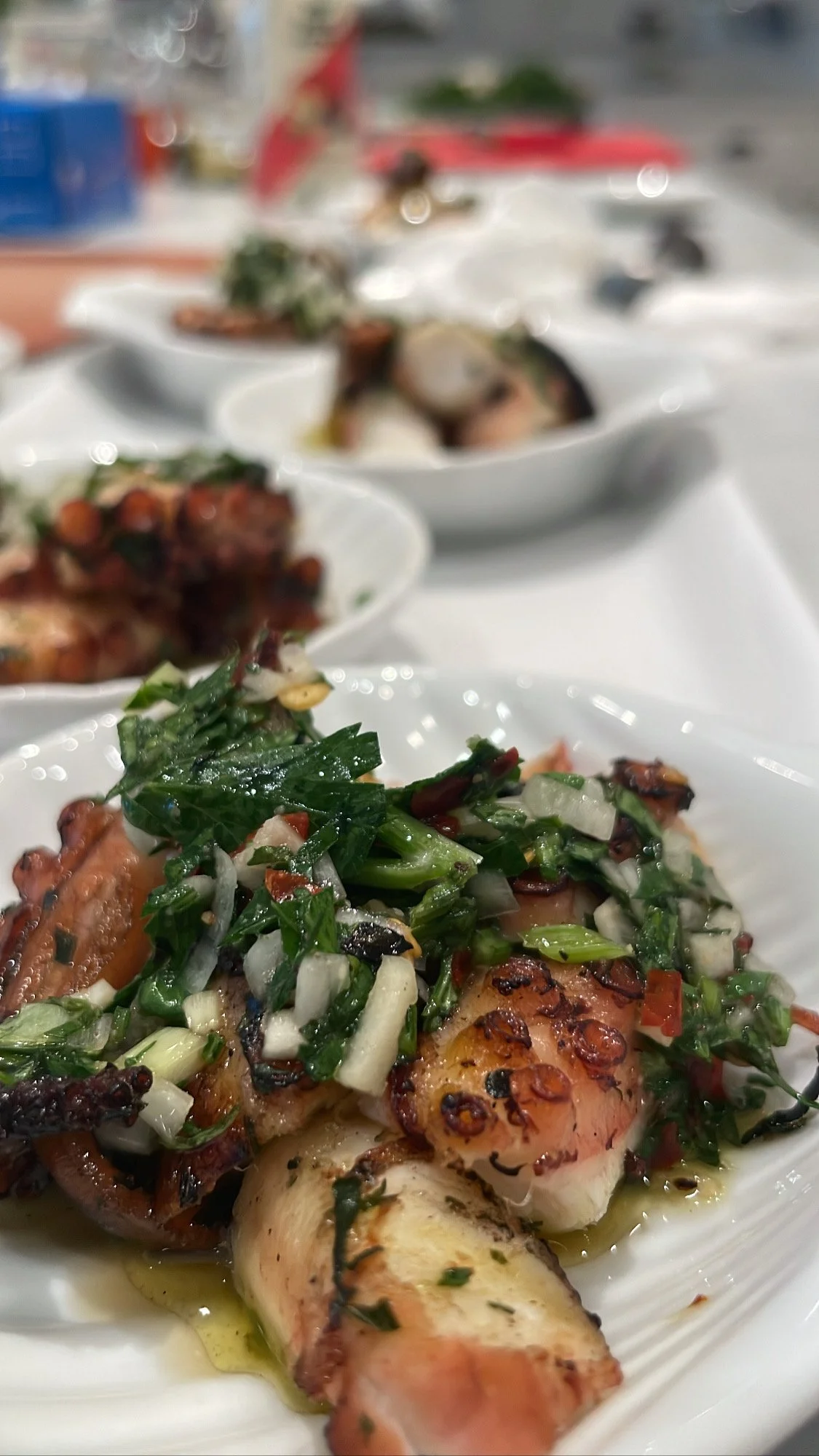 Close-up of grilled octopus topped with chopped herbs and vegetables on a white plate; other plates with seafood dishes blurred in the background.