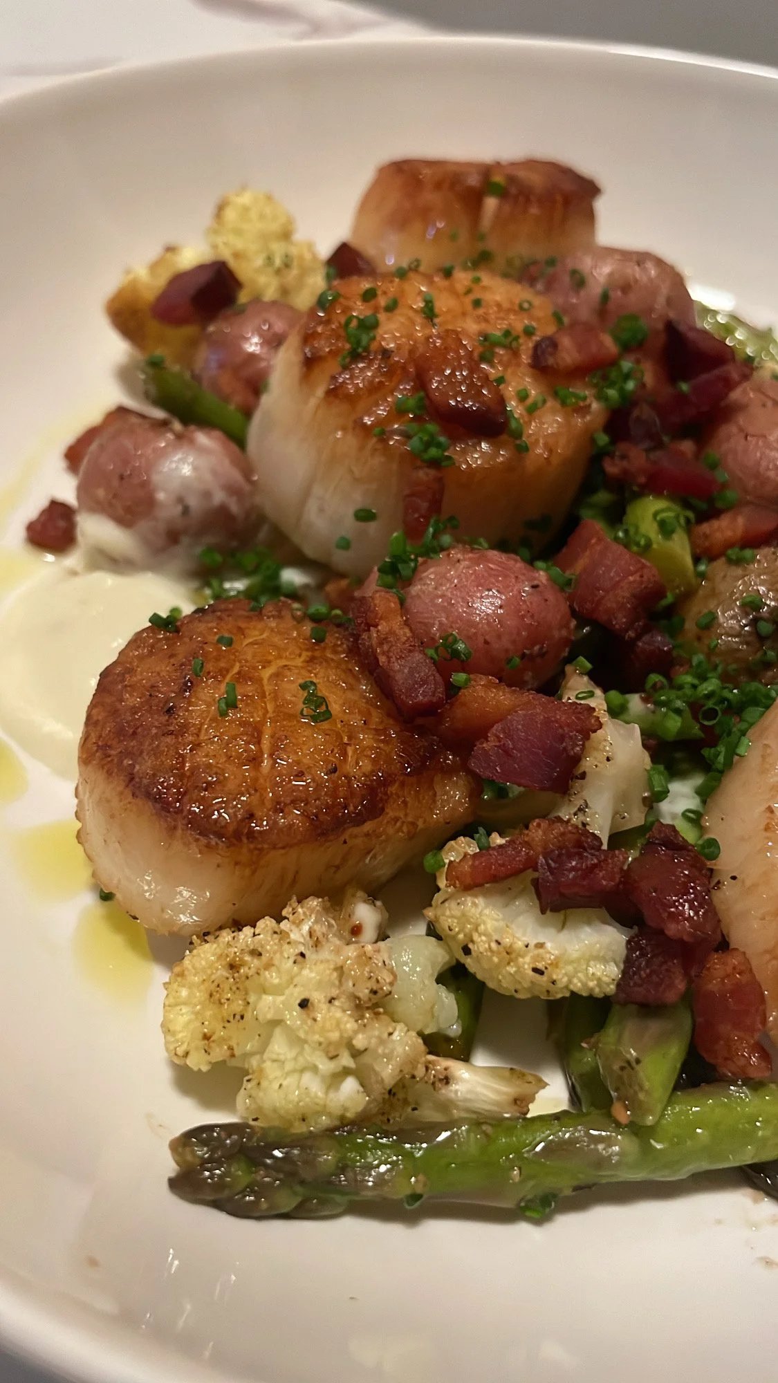 A plate of seared scallops with cauliflower, asparagus, bacon, and chorizo, garnished with chopped chives and drizzled with olive oil.