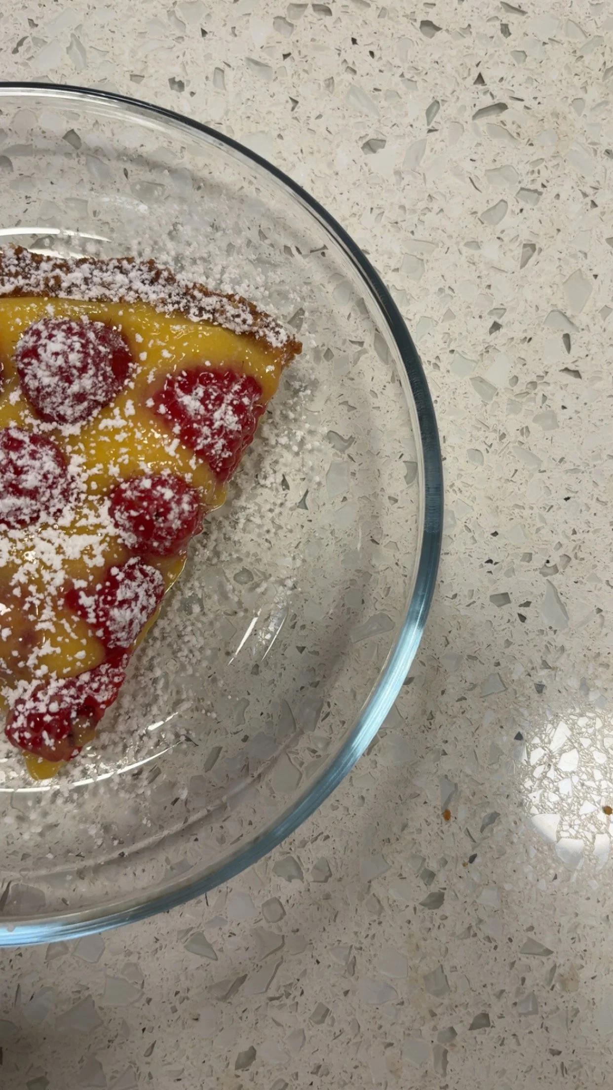 A slice of raspberry cheesecake with powdered sugar on top, in a glass dish on a speckled countertop.