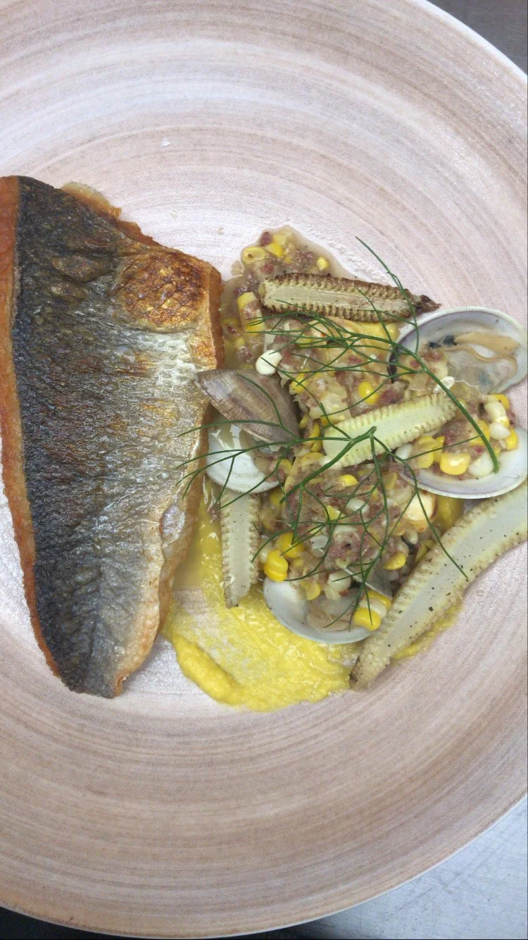 A plate with a cooked fish fillet, a bed of yellow sauce, and a seafood salad with clams, corn, and herbs.