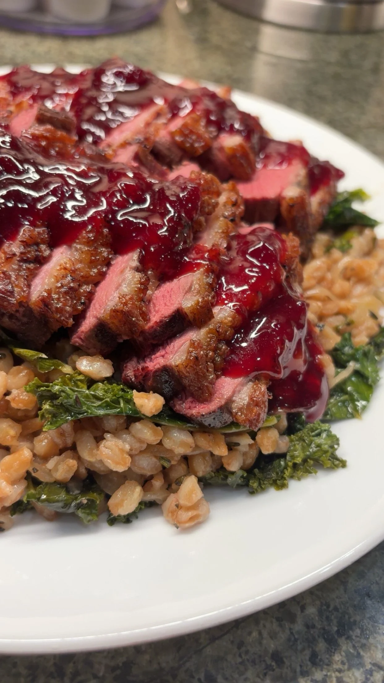 Sliced cooked steak topped with berry sauce, served on a bed of cooked farro and greens on a white plate.