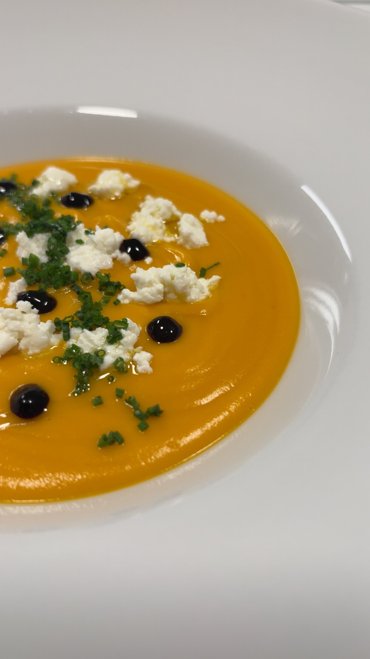 A bowl of orange-colored soup, garnished with crumbled white cheese, chopped chives, and black bubbles or caviar.