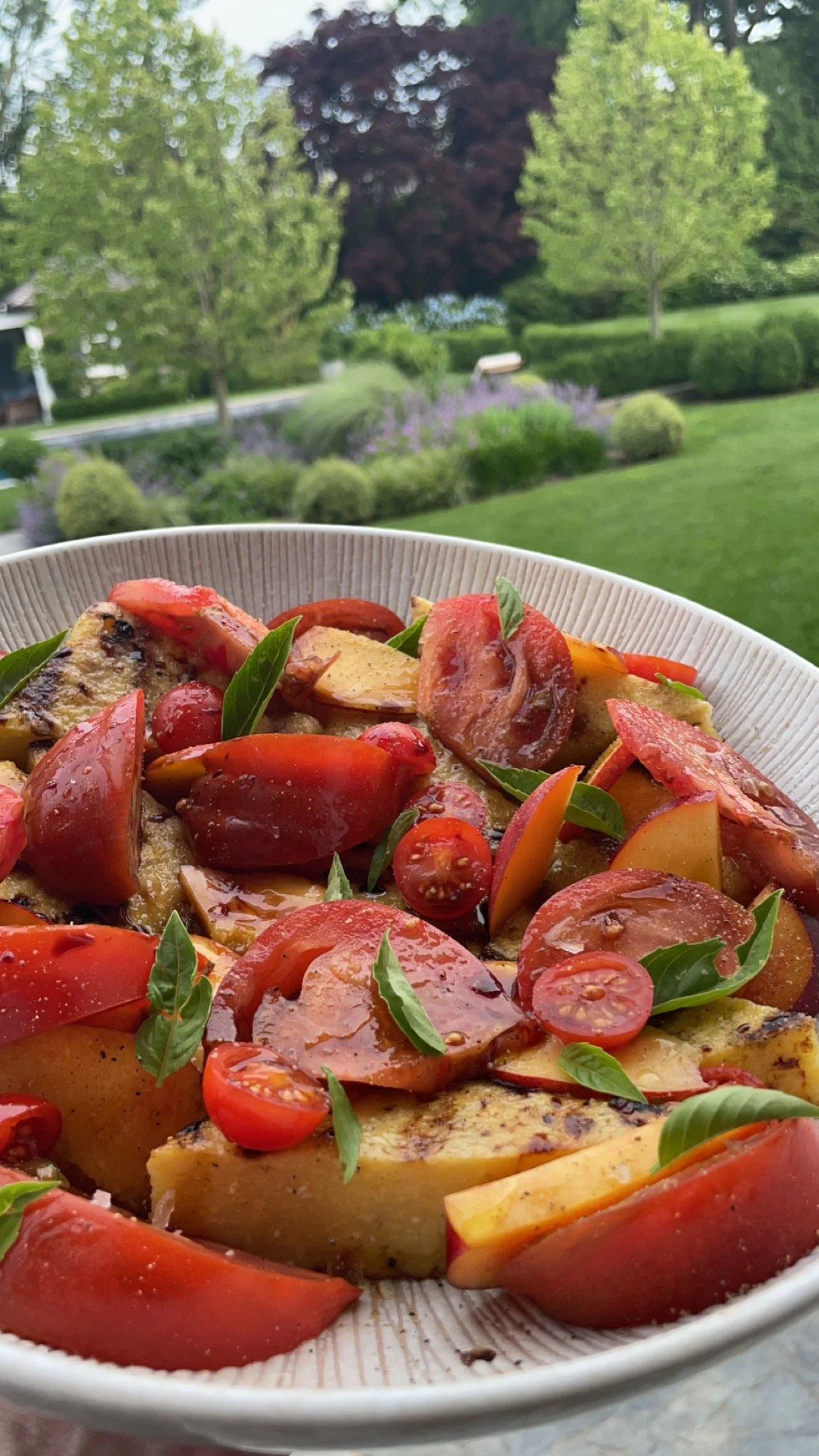 A bowl of salad with cherry tomatoes, apple slices, basil leaves, and grilled vegetables, set outdoors with a garden and trees in the background.