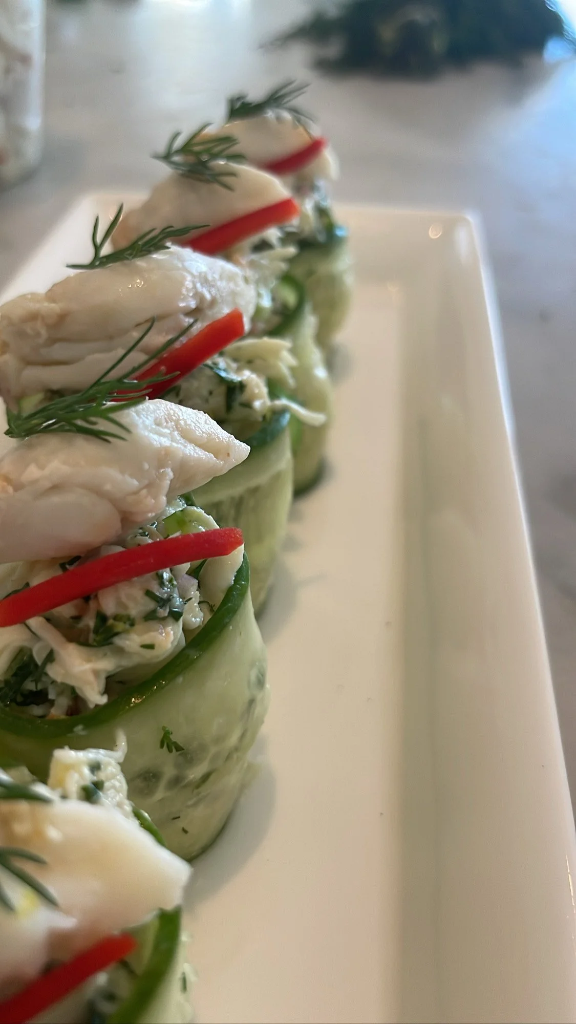 Rows of cucumber cups filled with seafood salad, garnished with red pepper strips and fresh herbs on a white rectangular plate.