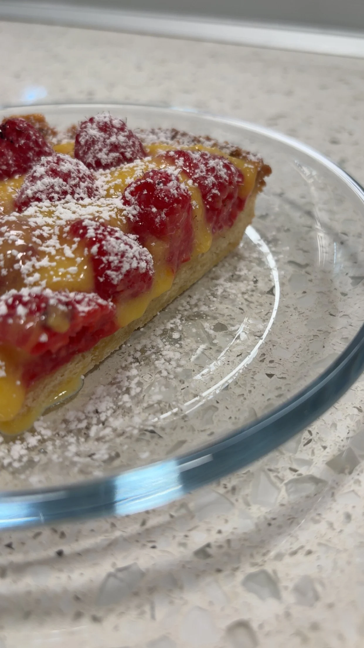 A slice of cherry tart with a lemon custard filling, topped with fresh cherries and powdered sugar, on a clear glass plate.