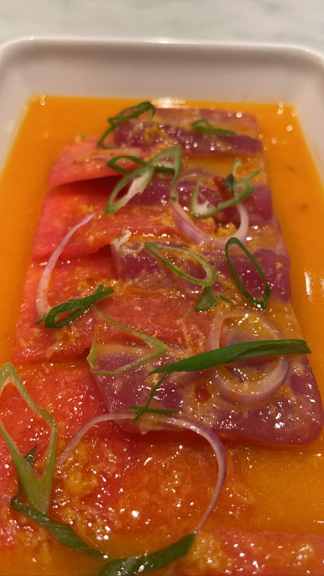 Close-up of sliced raw fish, possibly tuna, garnished with sliced red onions and chopped green onions, served in a light orange sauce or marinade.