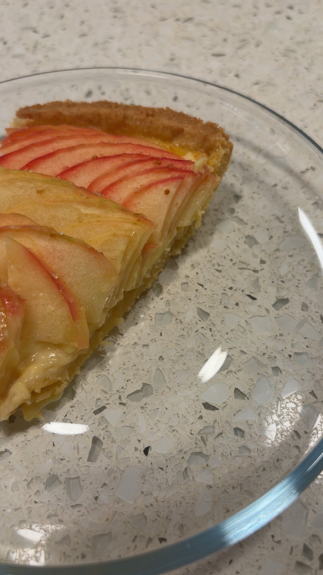 A slice of apple tart with thinly sliced apples on top in a glass dish on a speckled countertop.