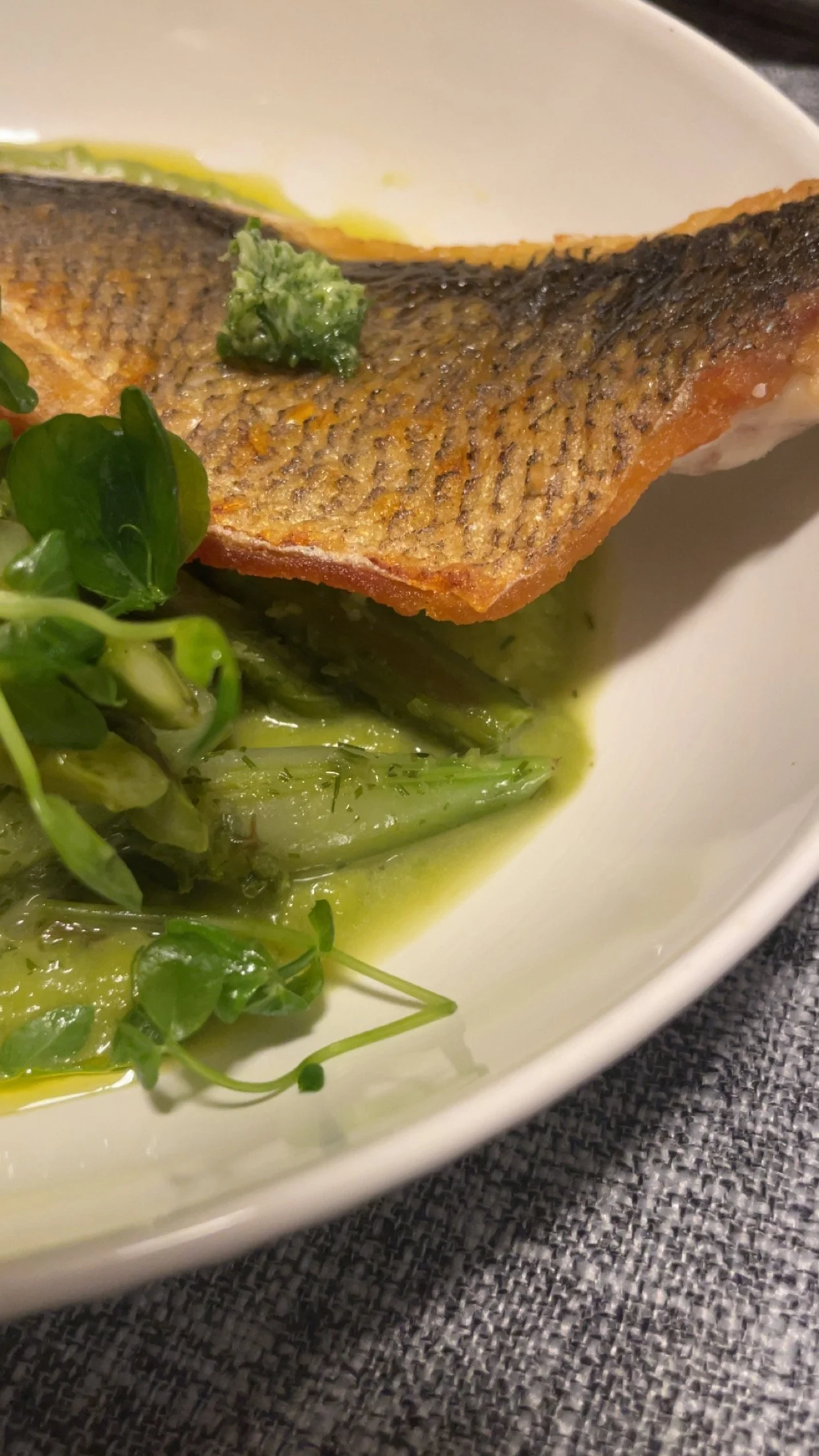 Close-up of a cooked fish fillet with crispy skin, served with a dollop of green wasabi, green microgreens, and green sauce or oil on a white plate.