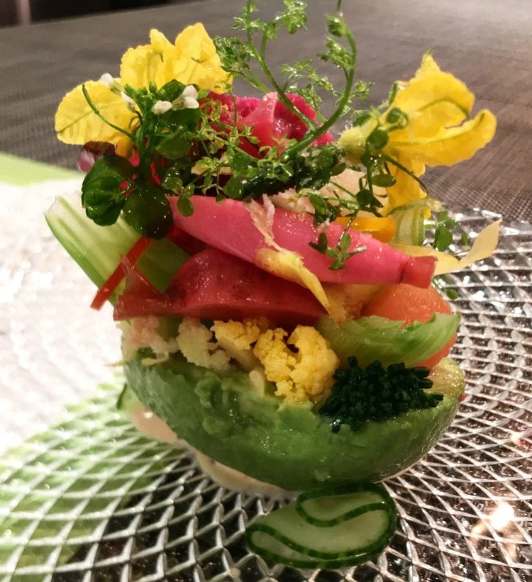 A colorful gourmet vegetable salad with various fresh vegetables, edible flowers, and greens on a textured glass plate.