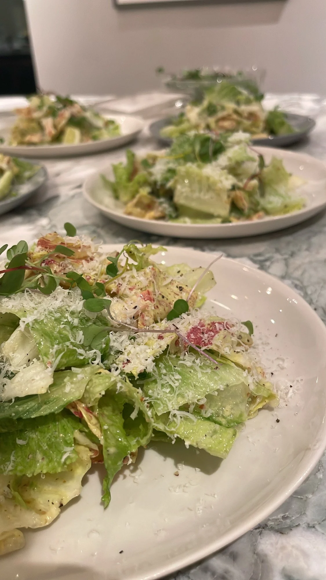 Close-up of a plate of Caesar salad with grated cheese and microgreens in the foreground, with multiple plates of similar salads on a marble table in the background.