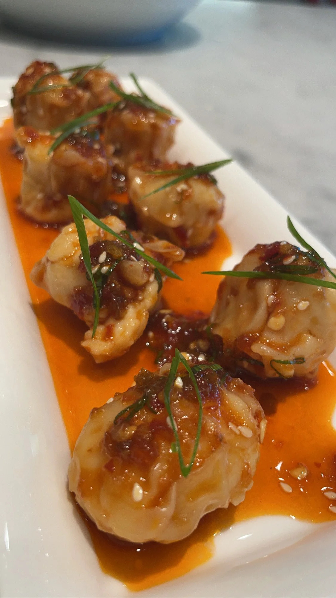 Close-up of six Asian-style dumplings topped with green herbs and sesame seeds, served on a white rectangular plate with orange sauce.