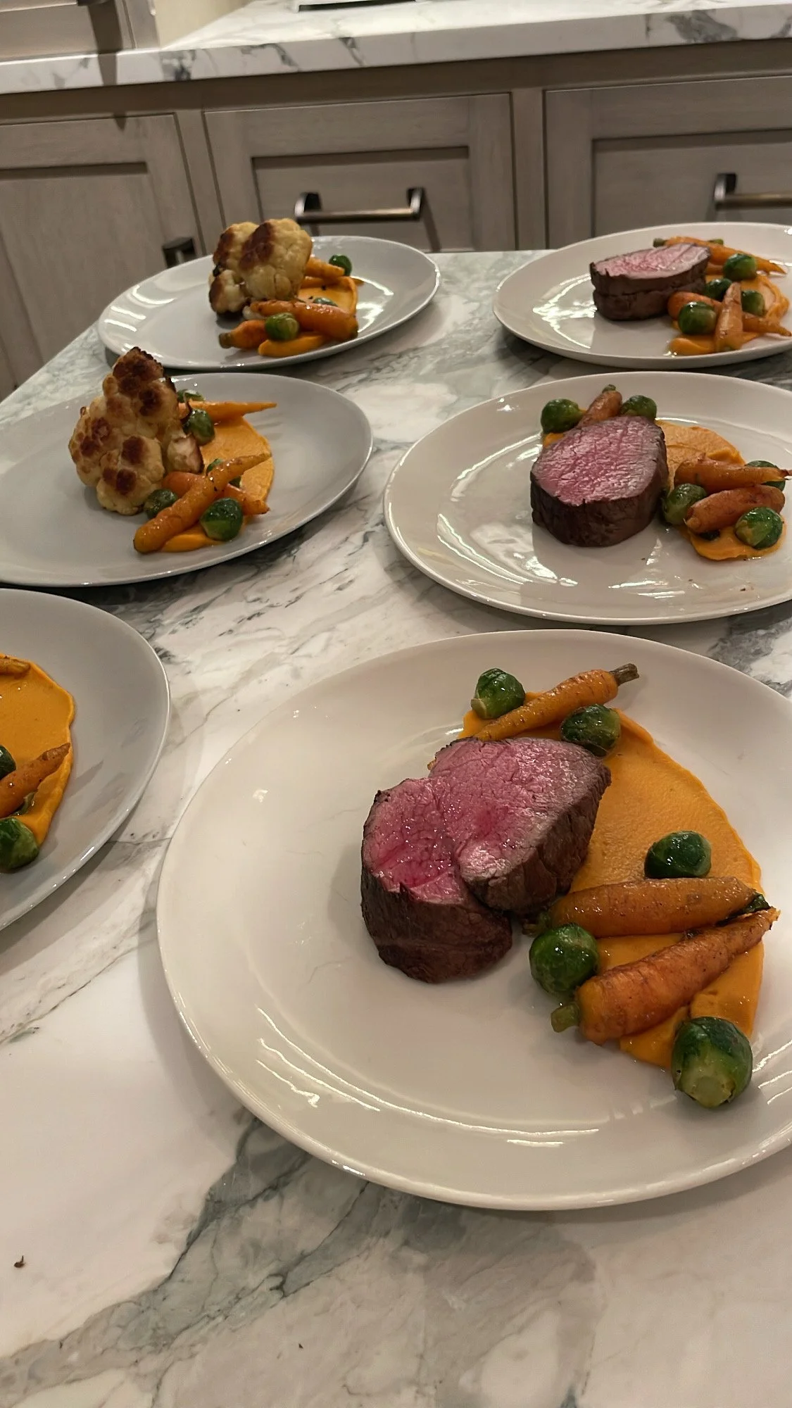 Plates with cooked beef tenderloin, roasted carrots, Brussels sprouts, and a smear of orange-colored puree, arranged on a marble countertop.