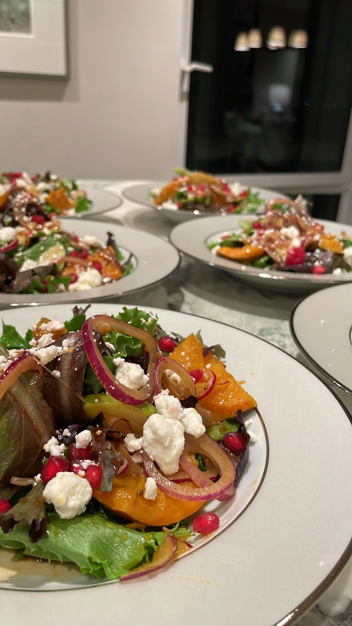 Multiple plates of colorful mixed salads with ingredients like pumpkin, red onions, pomegranate seeds, and cheese on a white table
