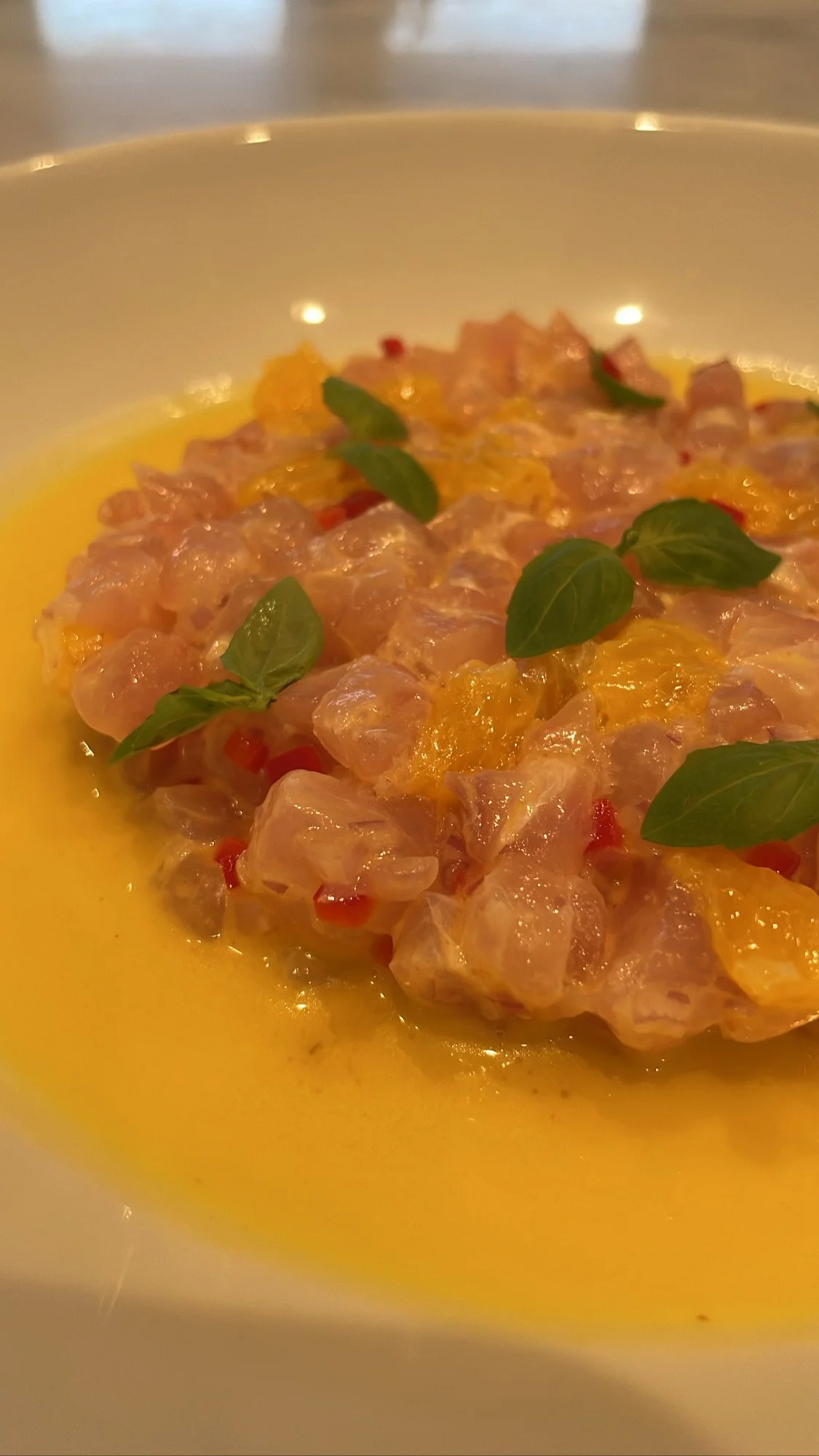 Close-up of a bowl containing finely chopped raw fish or seafood, garnished with fresh basil leaves, immersed in a yellow sauce or oil.