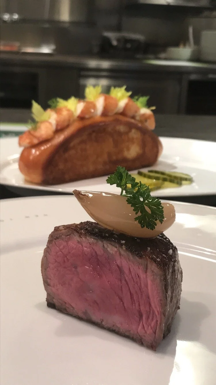 Close-up of a plate with a slice of cooked beef topped with a garlic clove and parsley, with a background of a hot dog with chili, mustard, relish, and a bun.