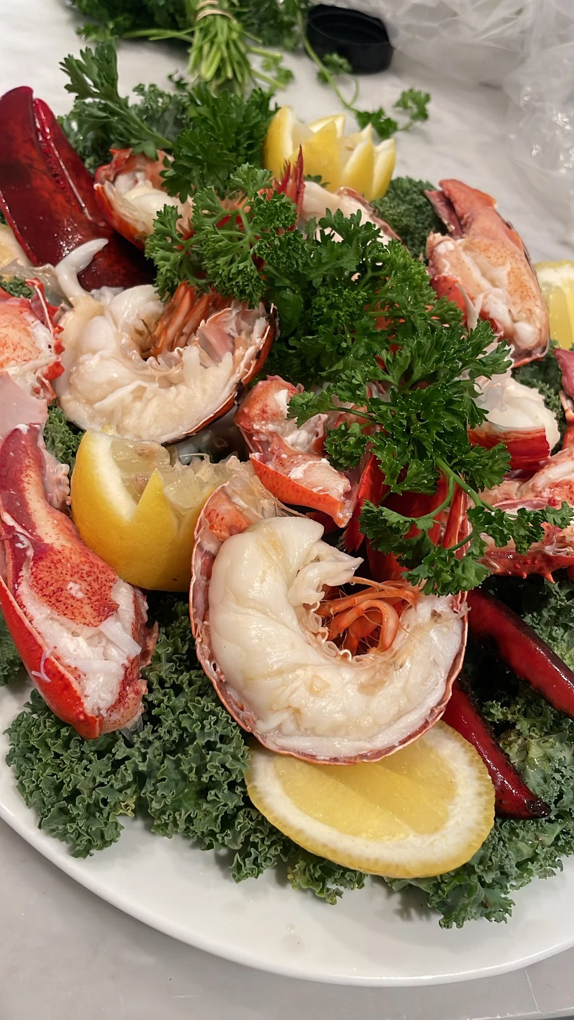 Seafood platter with lobster, shrimp, and garnishes including lemon wedges, parsley, kale, and hot peppers.