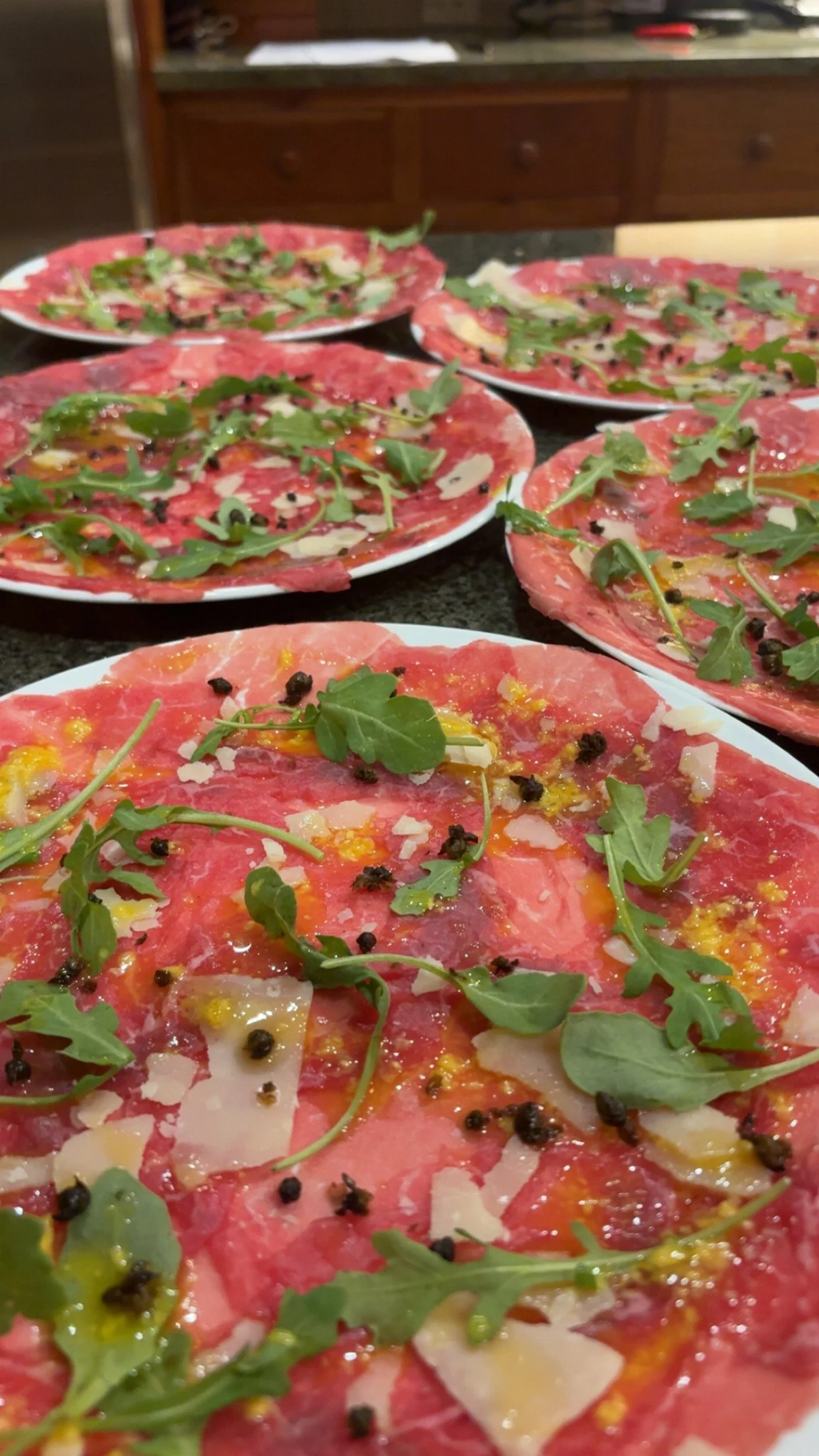 Several plates of carpaccio with arugula, shaved cheese, black pepper, and oil on a kitchen countertop.
