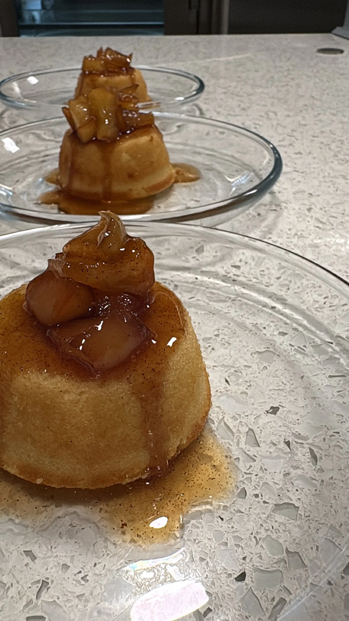 Three individual servings of caramel pudding topped with caramelized apple chunks on glass dessert plates, set on a white speckled surface.