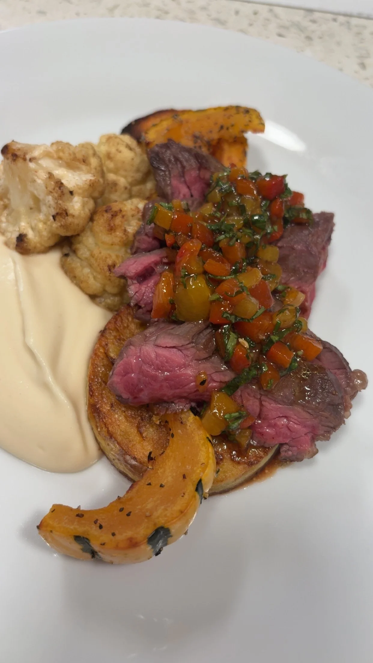Slices of cooked beef topped with a colorful pepper relish, accompanied by roasted pumpkin, cauliflower, and a dollop of creamy sauce on a white plate.