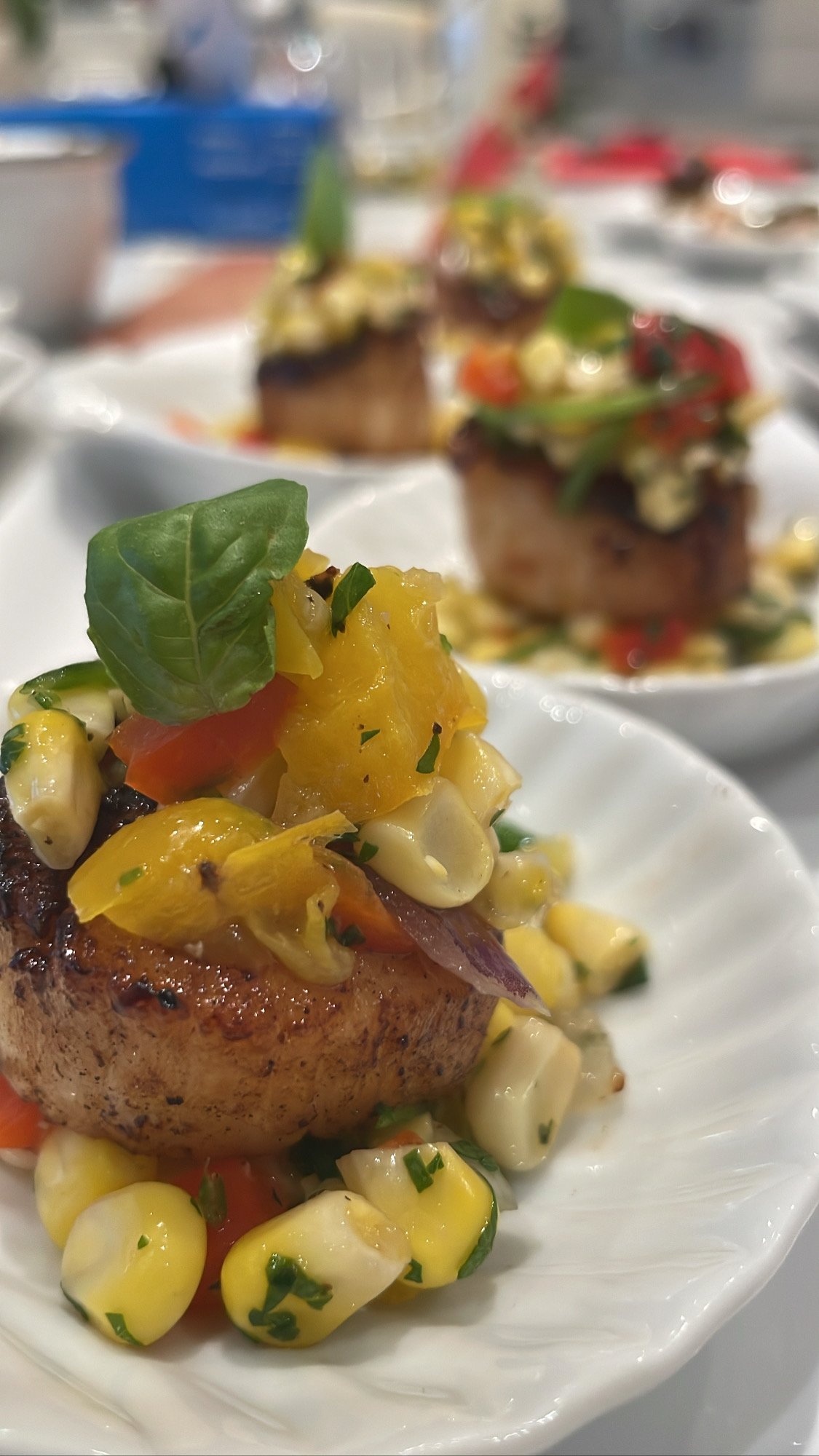 Close-up of a gourmet appetizer with grilled scallop topped with corn salad, garnished with basil, on a white plate.