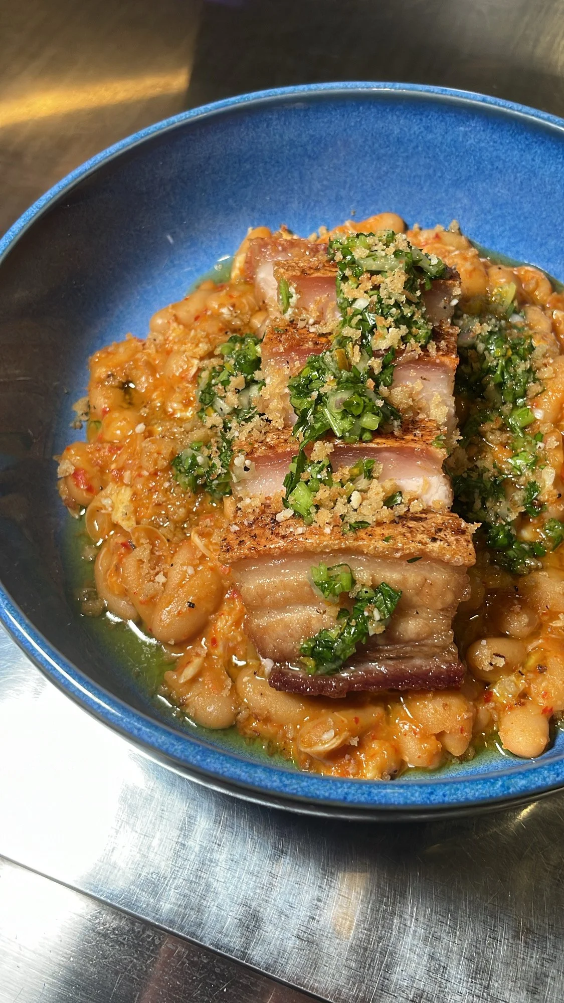 Close-up of a bowl of beans and roasted pork garnished with chopped green herbs and breadcrumbs.