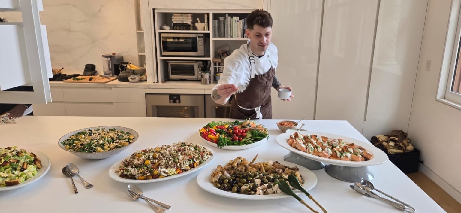 A man in a white shirt and brown apron stands behind a table filled with an assortment of colorful salads and dishes, in a bright kitchen with white cabinets and a window.