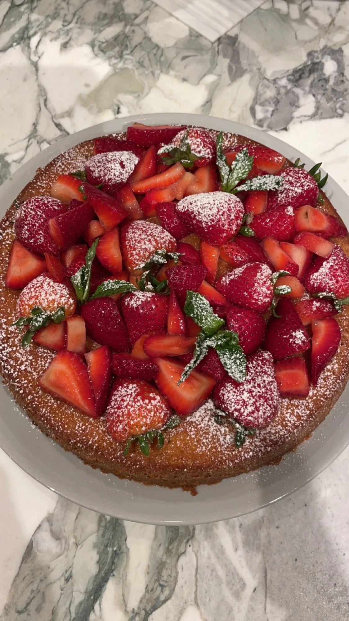 A fresh strawberry cake topped with sliced strawberries, powdered sugar, and mint leaves, on a white plate on a marble surface.