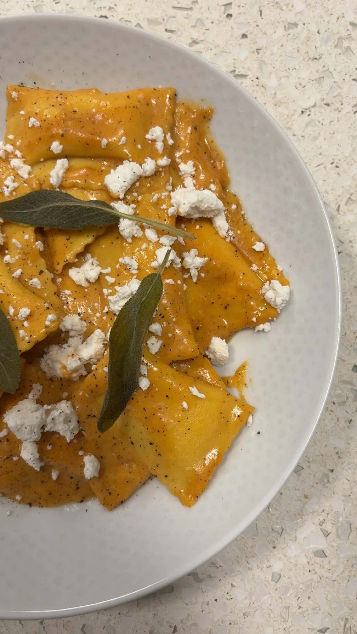 A white bowl of freshly cooked ravioli pasta topped with ricotta cheese, black pepper, and sage leaves on a light-colored countertop.