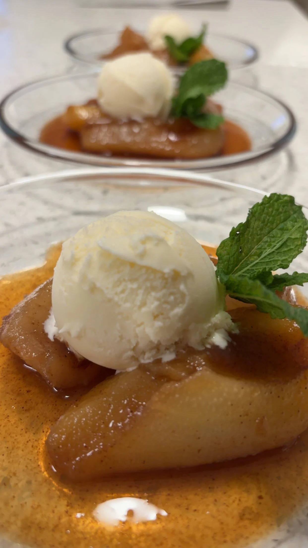 Close-up of a dessert with a scoop of vanilla ice cream, cinnamon-spiced sauce, a piece of fruit, and a mint leaf garnish in a glass dish.