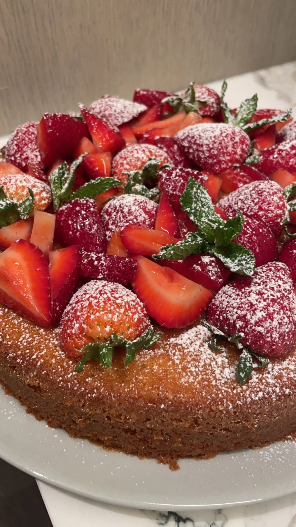 A round cake topped with sliced strawberries and fresh mint leaves, dusted with powdered sugar.