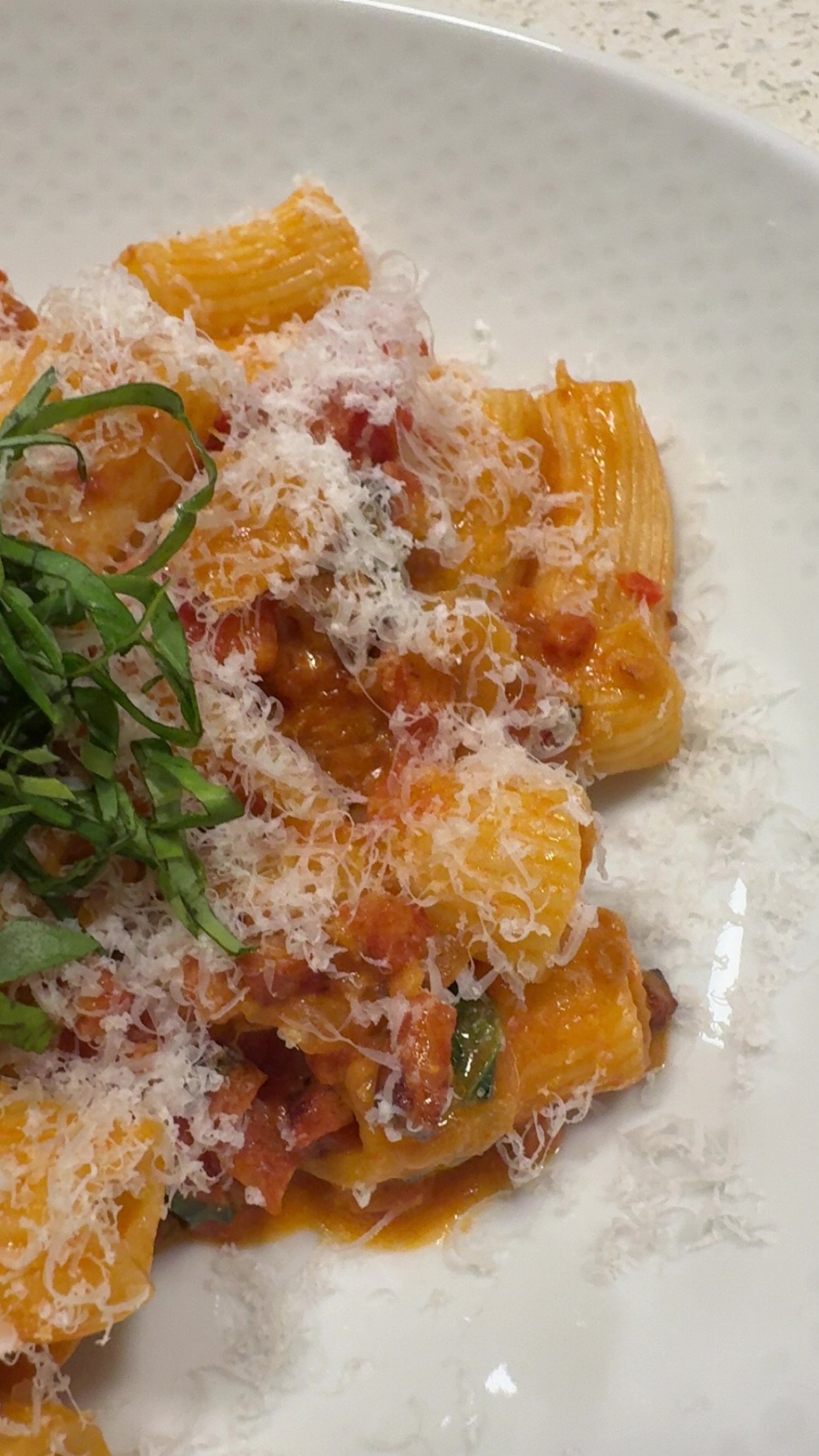 Close-up of cooked pasta with tomato sauce, grated cheese, and garnished with fresh herbs on a white plate.