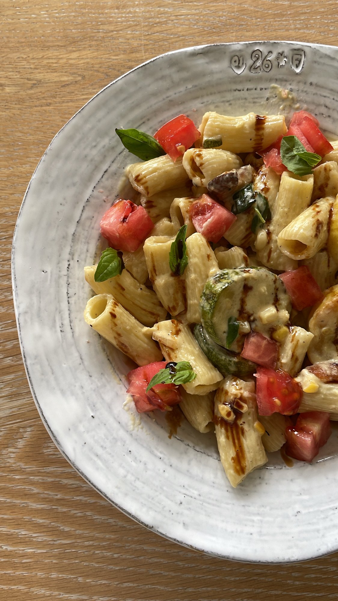 A plate of pasta with grilled zucchini, diced tomatoes, fresh basil leaves, and drizzled balsamic glaze on a wooden table.