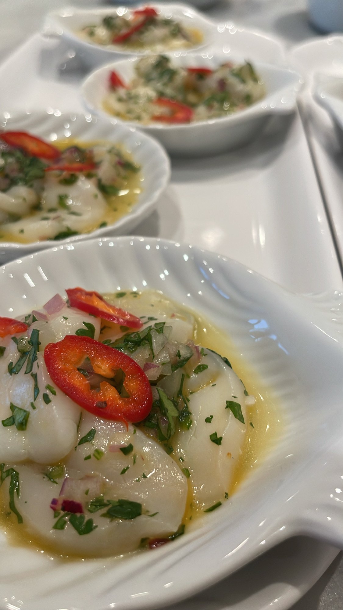 Close-up of ceviche served in white ceramic bowls, garnished with slices of red chili peppers, chopped onions, and herbs, with a lemon or lime citrus base.