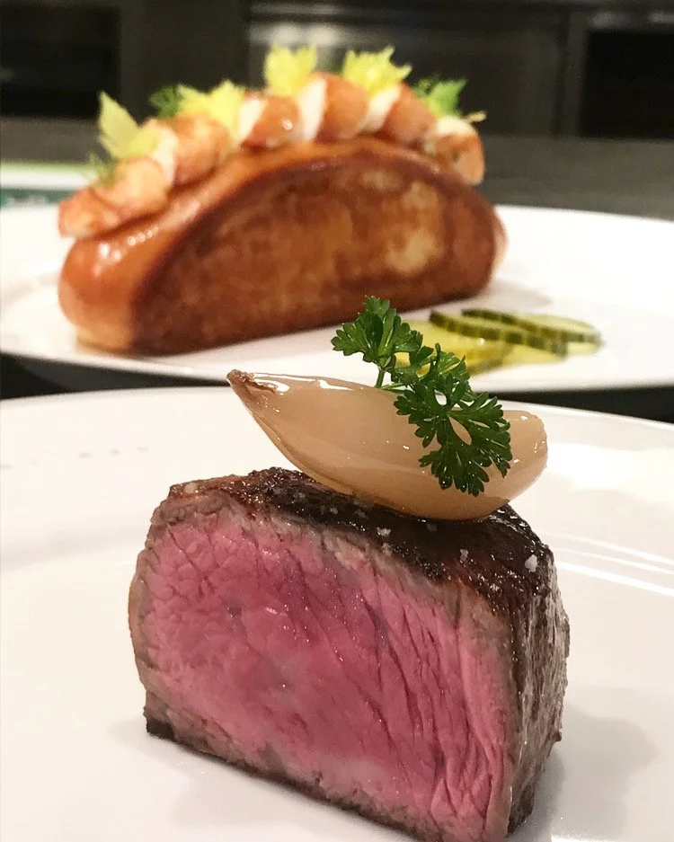 Close-up of a medium-rare steak topped with a dollop of sauce and a sprig of parsley, with a baked potato with toppings and a side of cucumber slices in the background.