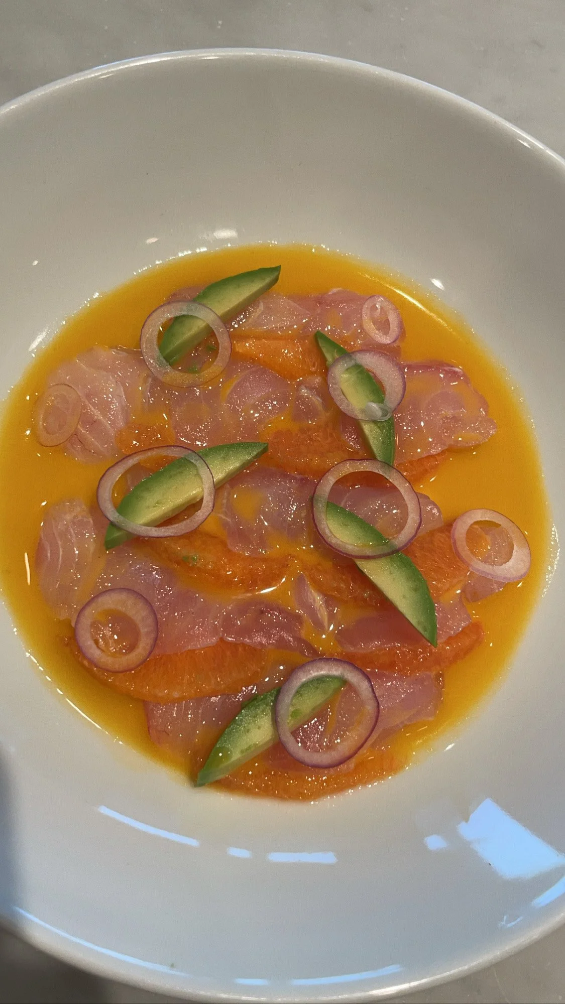 Slices of raw fish with sliced cucumbers, red onions, and orange sauce in a white bowl.