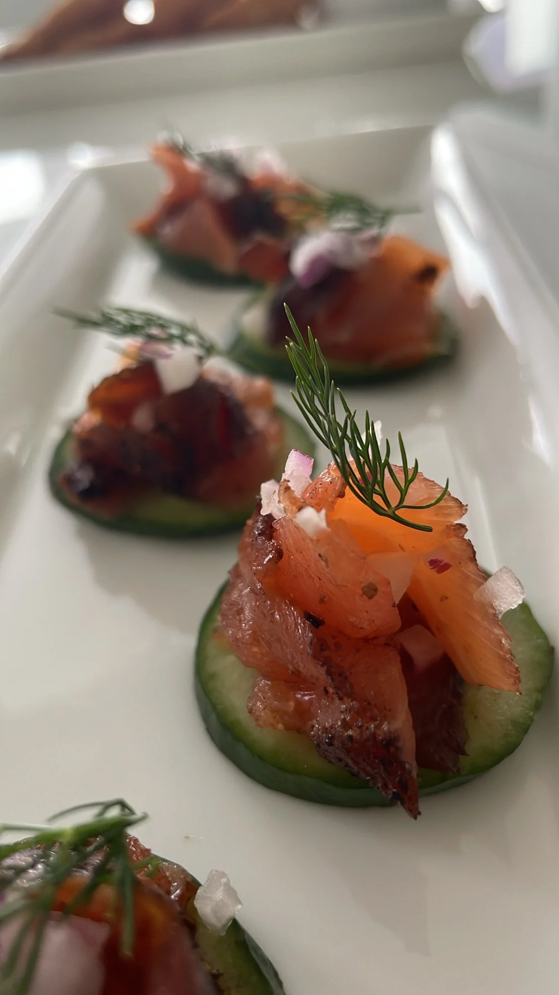 Close-up of smoked salmon atop cucumber slices, garnished with herbs, salt flakes, and edible flowers on a white rectangular plate.