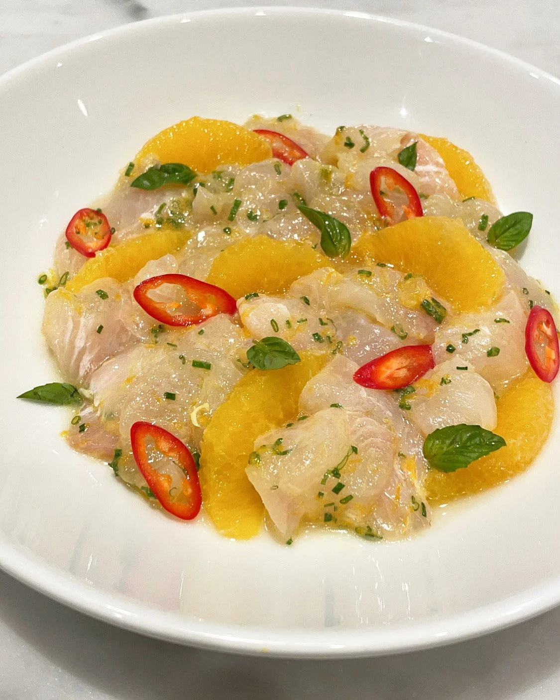 A plate of fish sashimi with orange slices, red chili pepper slices, and green herbs.