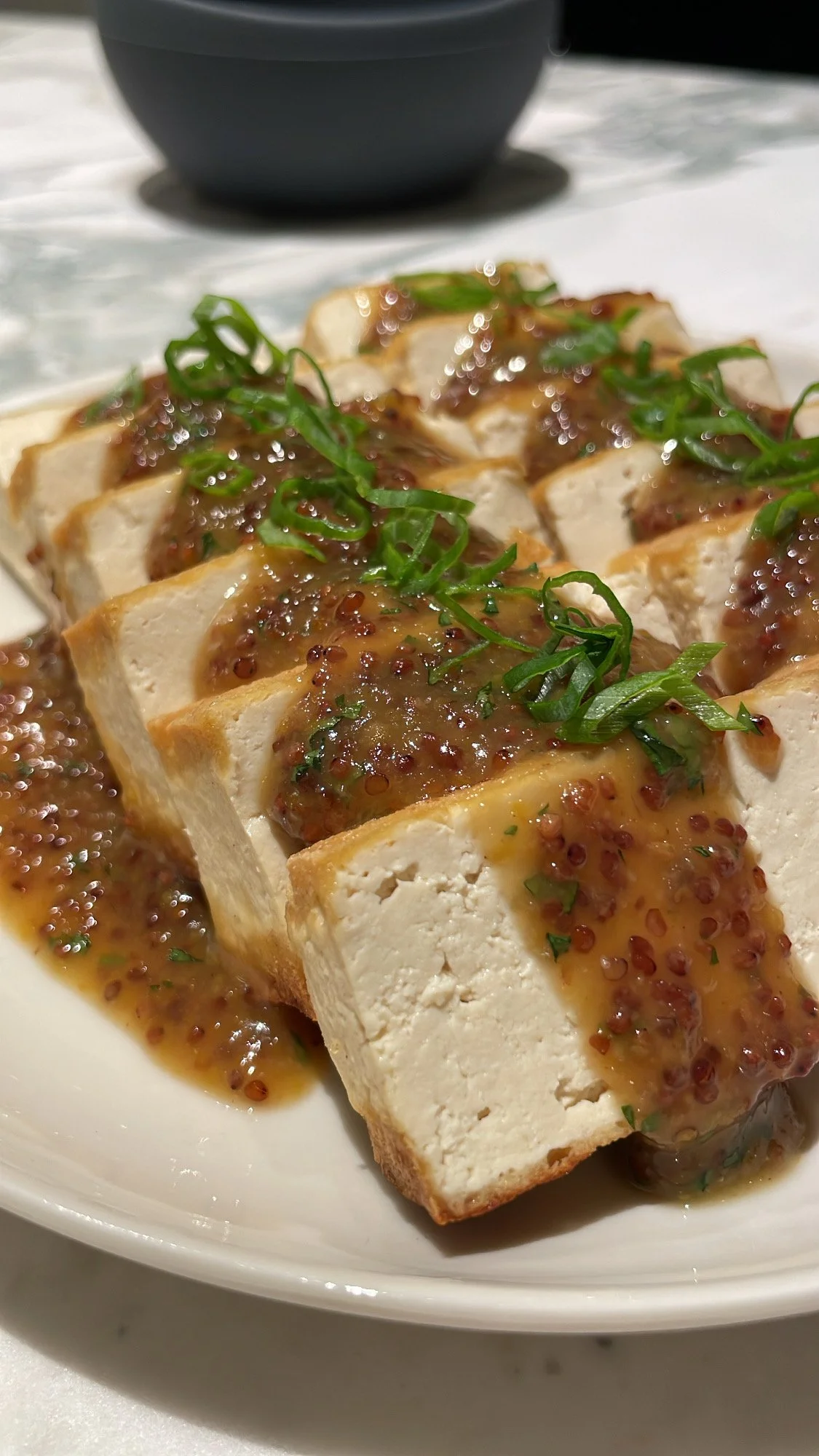 Tofu topped with mustard sauce and chopped green onions on a white plate.